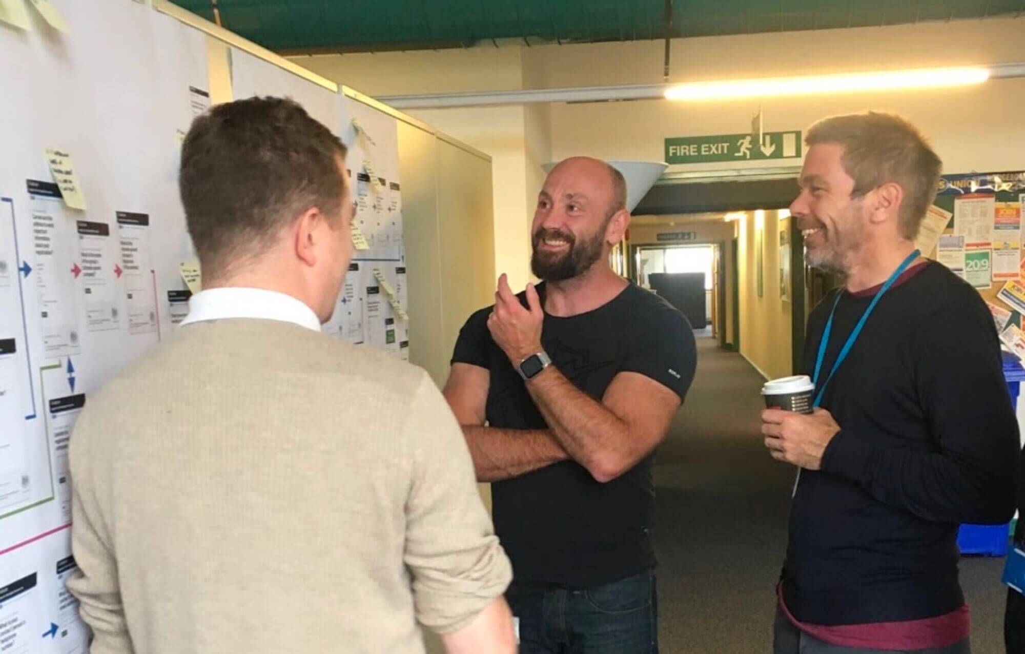 3 men stood close to a wall, discussing the contents of maps placed on a wall.